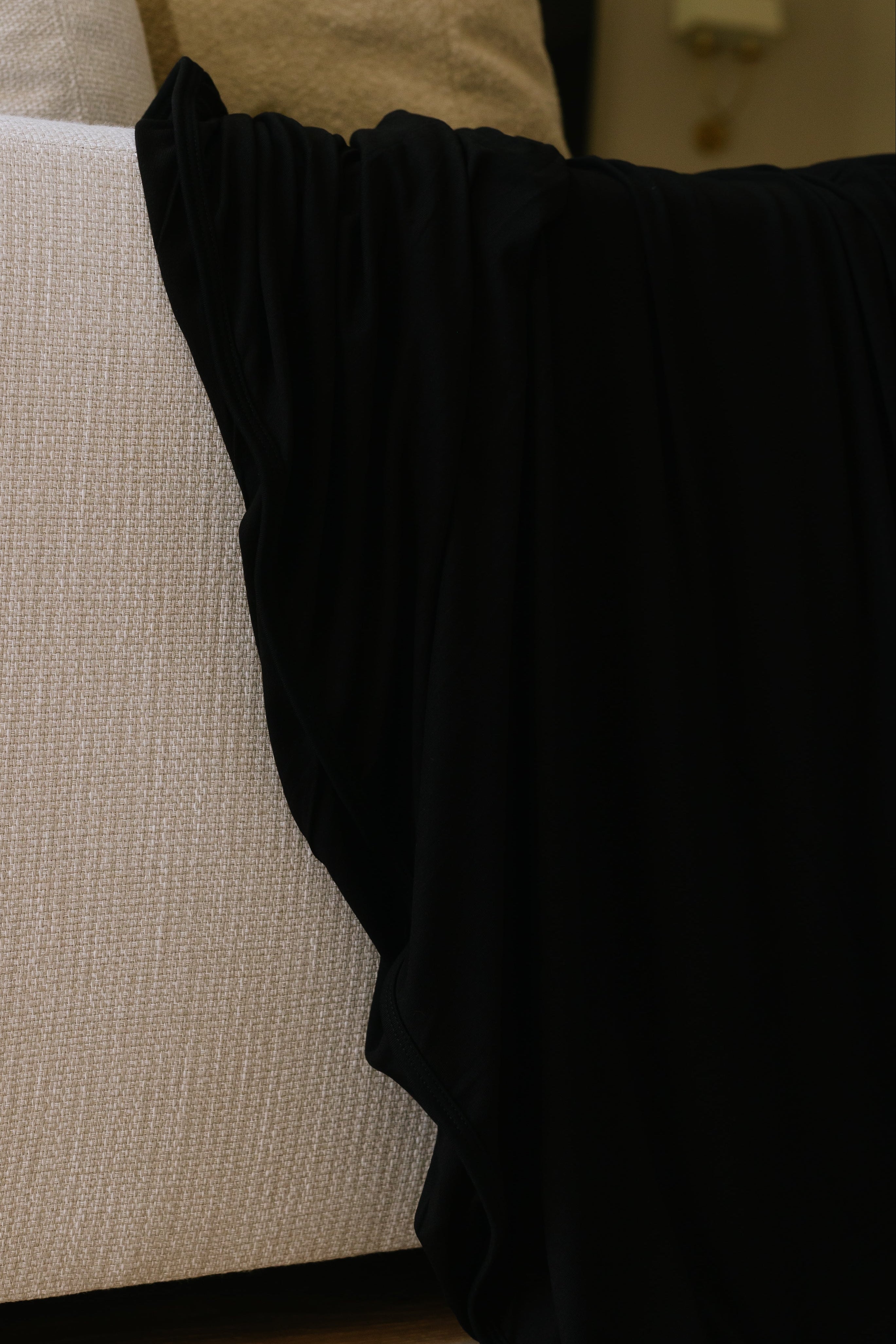 A close-up of the jet beige textured couch with cream pillows and a luxurious blanket draped over the side, set on a light wooden floor.