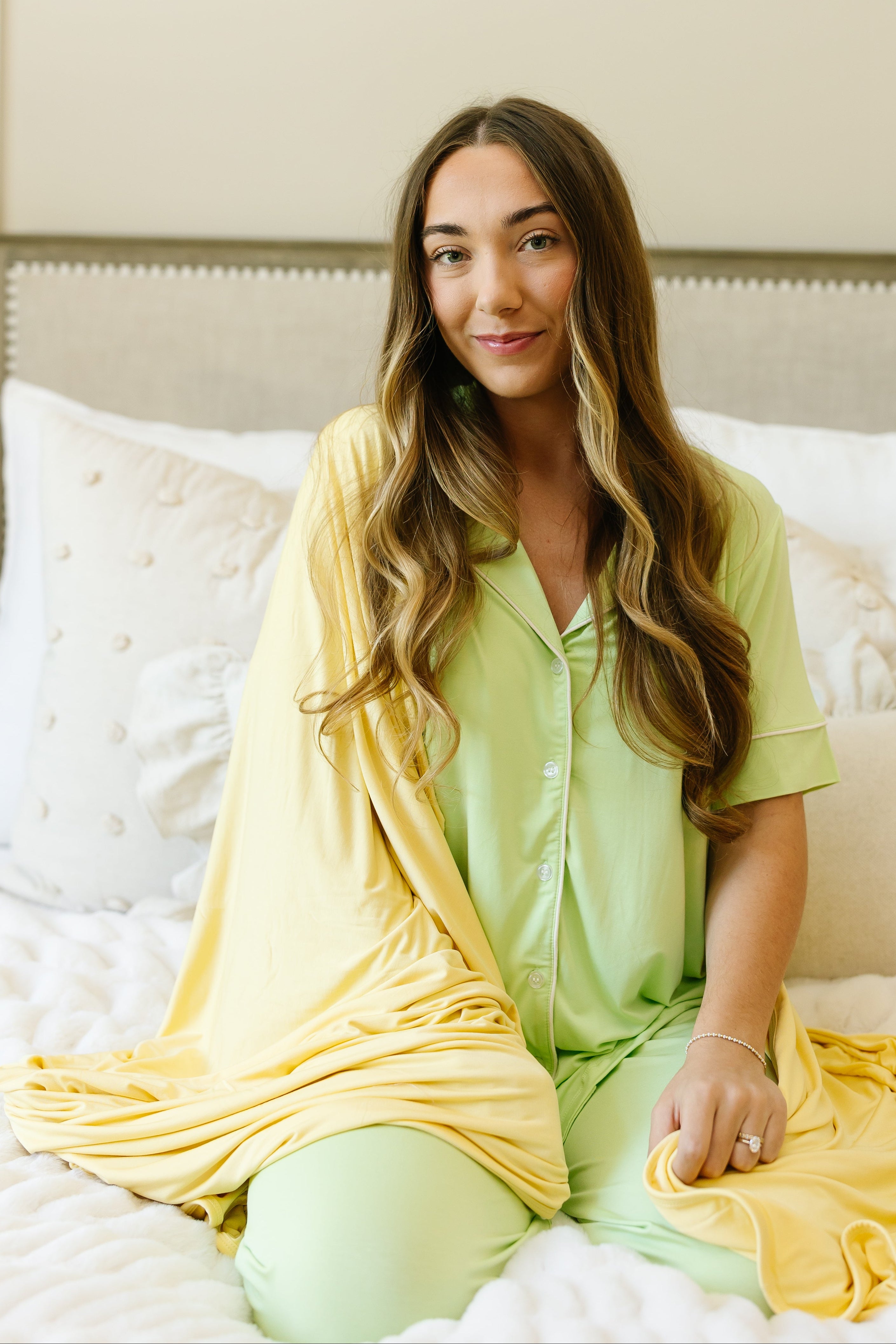 A woman with long brown hair smiles softly on a bed, wrapped in the lemon meringue modern blanket and wearing light green pajamas and a yellow robe, surrounded by white pillows and bedding in a bright, cozy room.