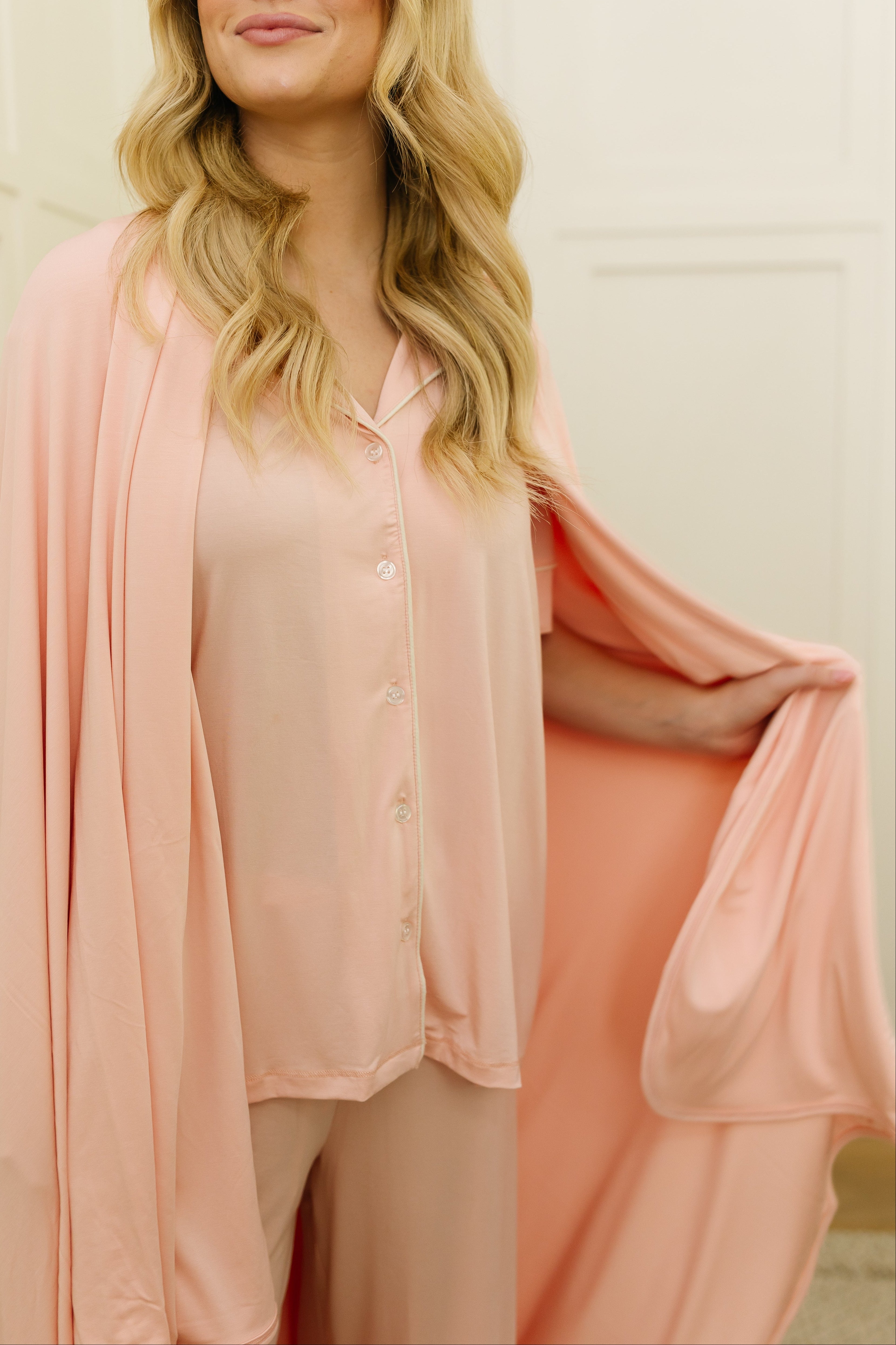 A woman with long blonde hair stands indoors against a cream wall, smiling to the side in a light pink outfit, elegantly draped with the modern “pinky” blanket over her shoulders.