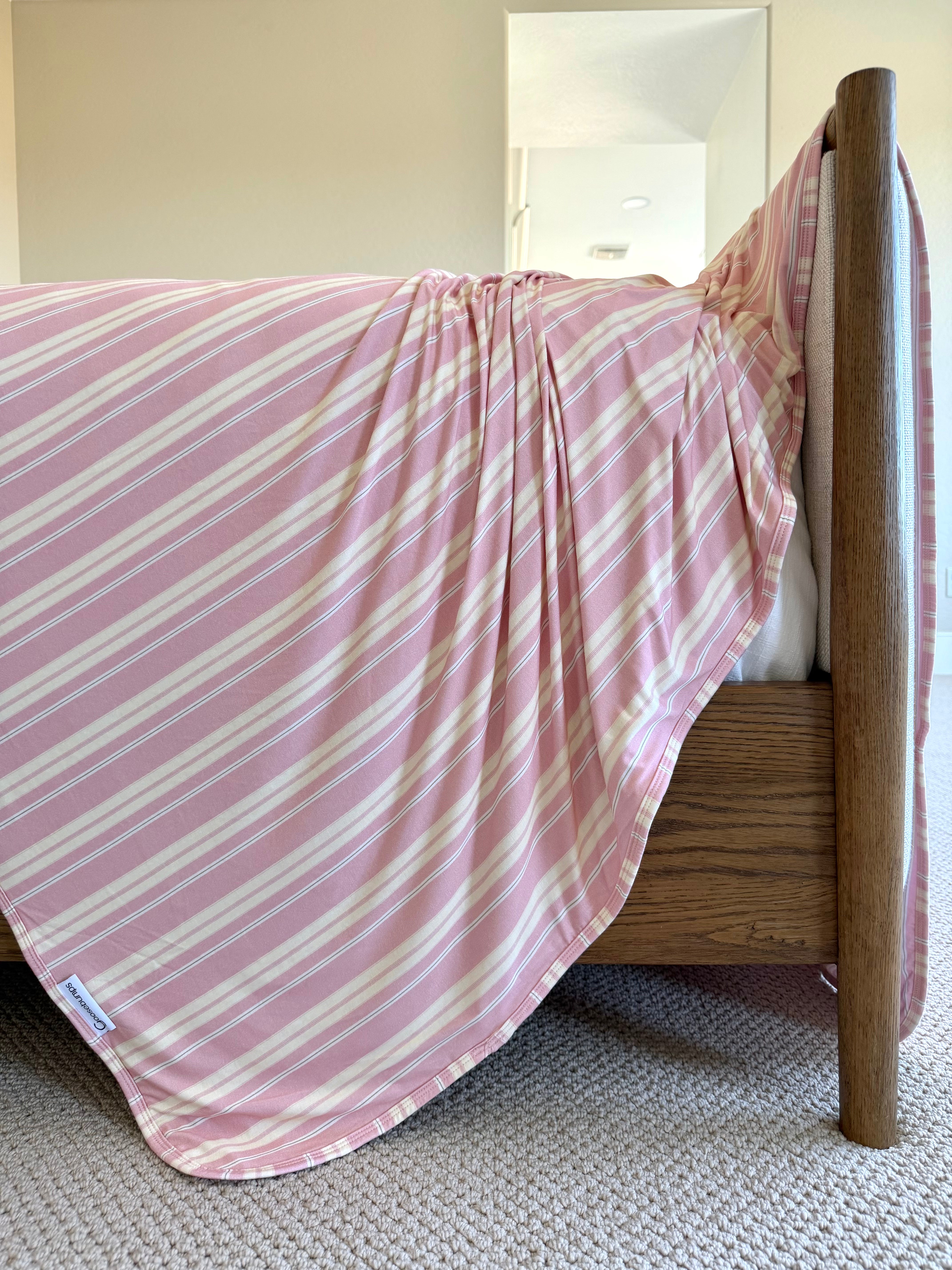 The Sherbet bamboo blanket, featuring pink and white stripes, is draped over a wooden bed frame in a softly lit bedroom with beige walls and carpeted flooring.