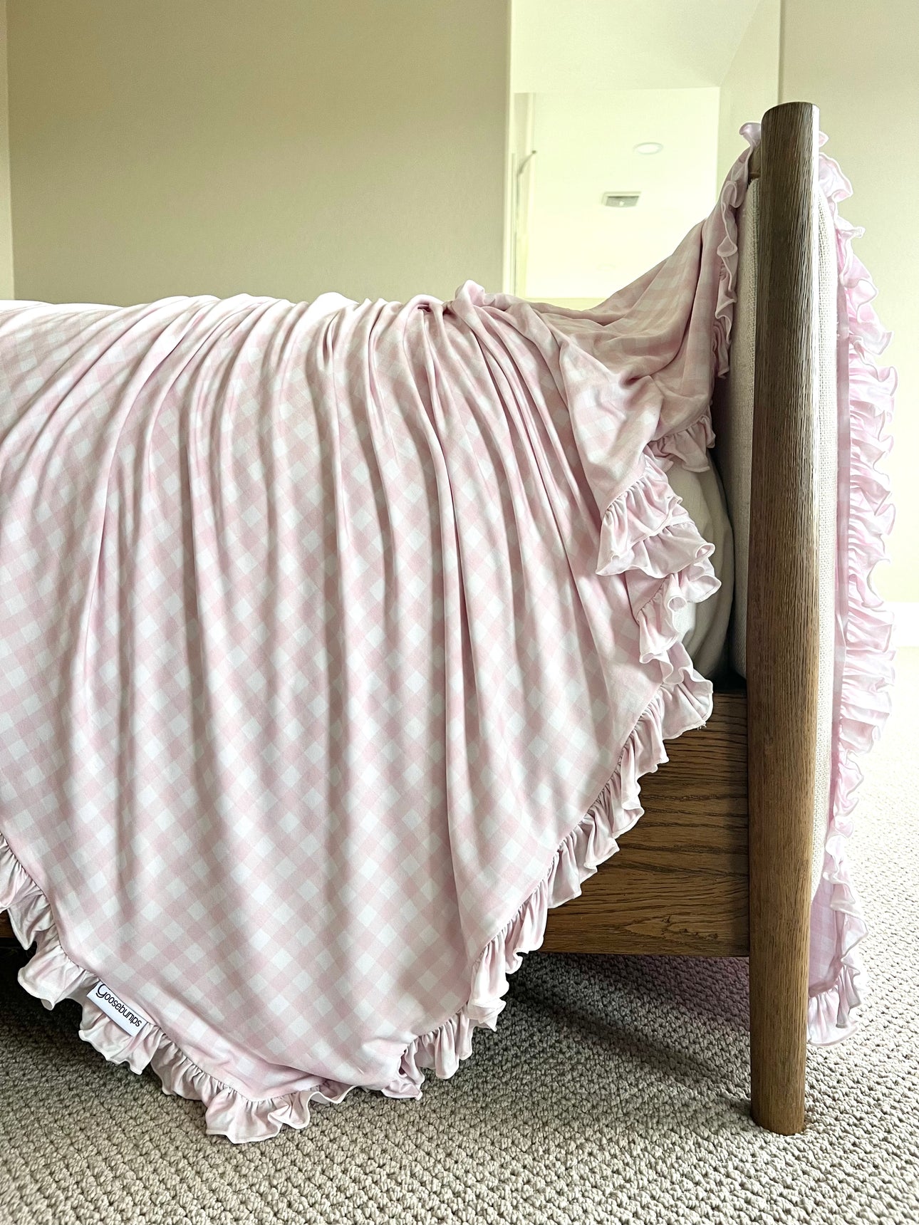 The xo / ruffle blanket in pink gingham, featuring delicate ruffled edges, is draped over a wooden footboard in a softly carpeted, neutral-toned room.